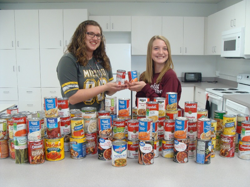 Two students standing behind dozens of cans of collected soup